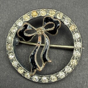 May include: A circular brooch featuring a dark metal frame set with clear and light yellow rhinestones. Centered within the circle is a black enamel bow motif with a metallic border. A horizontal pin mechanism is visible across the centre.
