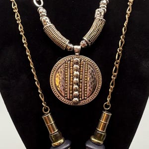 May include: A layered necklace set with gold and silver tones. The top necklace features a textured silver and gold chain. The middle necklace has a large, round pendant with a hammered texture. The bottom necklace has an elephant pendant.