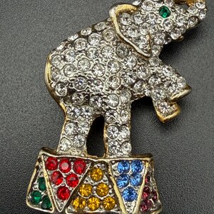 May include: A gold-tone metal brooch shaped like an elephant standing on a pedestal. The elephant is covered in clear rhinestones with a single green rhinestone eye. The pedestal features triangular sections filled with red, yellow, blue, and green rhinestones.