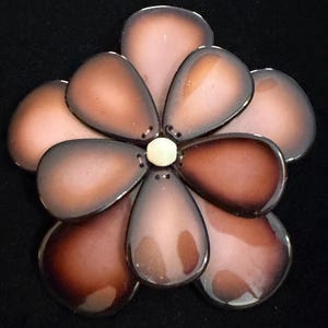 May include: A flower-shaped brooch with seven petals in shades of brown and pink, with a small, off-white center. The petals have a glossy finish and are arranged in a symmetrical pattern. The brooch is set against a black background.