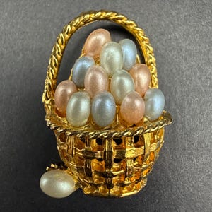May include: A gold-tone metal brooch shaped like a woven basket with a twisted handle. The basket is filled with multiple small, oval-shaped faux pearls in soft shades of peach, pale blue, and cream, with one additional pearl resting at the base.