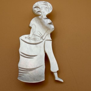 May include: A silver-colored brooch depicting a person playing a drum. The figure is wearing a hat and pants, and is holding drumsticks. The brooch is set against a brown background.