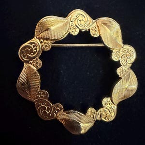 May include: A gold-toned brooch with a circular design. The brooch features leaf and swirl details around the perimeter, with a pin closure on the back. The jewelry piece is set against a dark background.