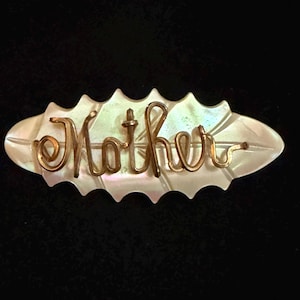 May include: A vintage brooch with the word "Mother" in gold script. The word is set on a pearlescent, oval-shaped base with a scalloped edge. The background is black.