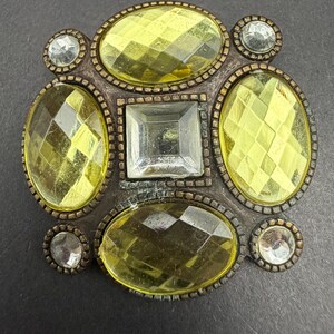 May include: A decorative brooch featuring a central square clear stone surrounded by four larger oval yellow faceted stones and four smaller round clear stones, all set within a dark metallic frame with a textured, beaded border design.