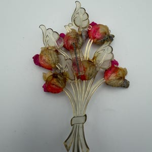 Könnte beinhalten: Eine klare, durchscheinende Brosche in Form eines Blumenstraußes, der mit einem Band zusammengebunden ist. Das Design zeigt geformte Kunststoffstiele und Blätter mit eingebetteten, texturierten, kugelförmigen Elementen in Rosa, Rot und Beige.