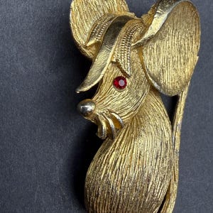 May include: A gold-toned metal brooch shaped like a mouse with a textured, brushed finish. The mouse features a single small, round red rhinestone for an eye, a smooth metallic nose, and a curved tail that extends along the side of the body.