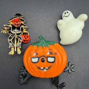 May include: Three Halloween-themed pins: a gold-tone scarecrow with a pumpkin head and bucket, a white translucent ghost with a smiling face, and a bright orange pumpkin character wearing black-rimmed glasses with black arms and legs.