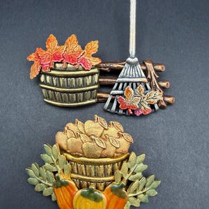 May include: Two metal brooches featuring autumn themes. The top brooch depicts a wooden barrel with orange and red maple leaves next to a metal rake. The bottom brooch shows a basket filled with apples, surrounded by green leaves and three orange pumpkins.