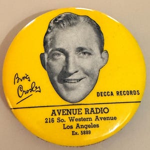 May include: A vintage yellow button featuring a black and white portrait of Bing Crosby. The button includes the text "DECCA RECORDS" and "AVENUE RADIO, 216 So. Western Avenue, Los Angeles, Ex. 5889". Bing Crosby's signature is also present.