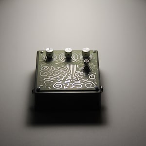 Könnte beinhalten: Ein grünes Metall-Effektpedal mit drei Knöpfen und einem Schalter. Das Pedal hat ein weißes Design mit einem wirbelnden Muster.