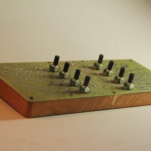 May include: A rectangular electronic device with a wooden base and a green top. The green top features a circuit board design and eight black knobs. It is likely a synthesizer or audio control module.