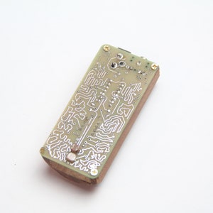 Puede incluir: Primer plano de la parte posterior de una placa de circuito con un revestimiento de epoxi verde y trazas plateadas. La placa está montada sobre una base de madera.