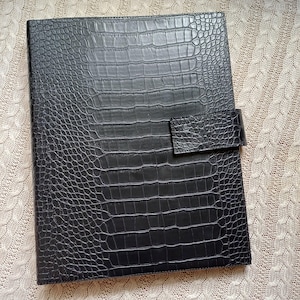 Puede incluir: Carpeta de cuero sintético negro con textura de piel de cocodrilo. La carpeta tiene forma rectangular con cierre de solapa. La cubierta es de color negro liso y la textura le da un aspecto sofisticado.