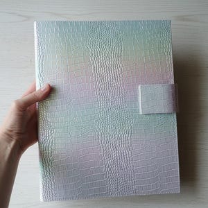 May include: A large, iridescent binder with a crocodile skin texture. The cover transitions between pastel colors, including pink, blue, and green. A rectangular clasp secures the binder. The binder is held by a hand.