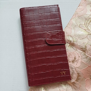 Puede incluir: Una cartera de cuero burdeos con textura de piel de cocodrilo. La cartera tiene un cierre de solapa y un monograma "VY" dorado. La cartera es rectangular y parece estar cerrada. El fondo es una tela con estampado floral.