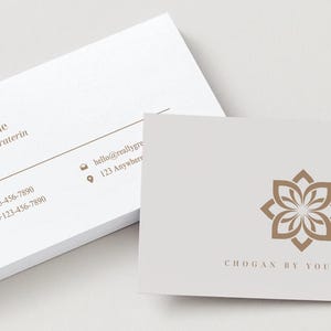Op de afbeelding: Witte visitekaartjes met bruine tekst en een bloemenlogo. Eén kaart toont contactgegevens, waaronder een telefoonnummer en e-mailadres. De andere kaart toont het logo en de tekst "CHOGAN BY YOUR NAME."