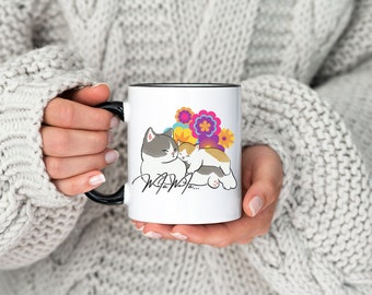 Könnte beinhalten: Weiße Keramiktasse mit schwarzem Rand, mit einer Cartoon-Illustration von zwei Katzen, die sich inmitten bunter Blumen kuscheln. Der Text "Meow" ist unten in Kursivschrift geschrieben.