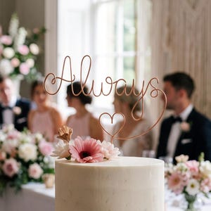 Op de afbeelding: Een tweelaagse wit geglazuurde taart met een koperkleurige draad-taarttopper die het woord Always in cursief schrift met een klein hartje vormt. De taart is versierd met roze en witte bloemen aan de basis van de topper en op de onderste laag.