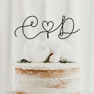 Może przedstawiać: Biały tort ślubny z czarnym drucianym topperem, na którym widnieje napis "C & D" z sercem w środku. Tort jest ozdobiony białymi piwoniiami i zielonymi liśćmi.