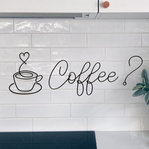 May include: White kitchen cabinets with a white tiled backsplash. A black wire sculpture of a coffee cup with a heart-shaped steam rising from it and the text "Coffee?" is on the backsplash. A vase with a pink protea flower arrangement is on the counter.