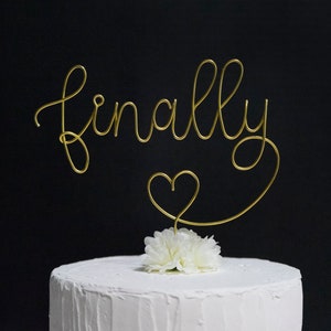 May include: Gold wire cake topper with the word "finally" and a heart. The topper is on a white cake with a white flower.