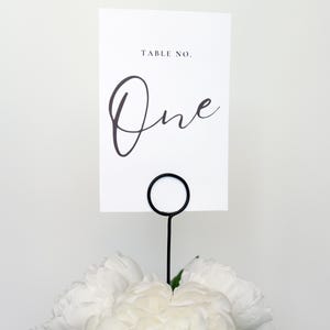 Peut inclure: Une carte de numéro de table blanche avec le texte « TABLE NO. » et le mot « One » en écriture élégante. La carte est tenue par un support en fil noir au-dessus d'un bouquet de pivoines blanches.