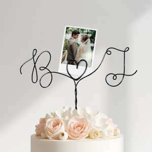 Könnte beinhalten: Ein schwarzer Draht-Kuchenaufsatz mit den Buchstaben "B" und "J" und einem herzförmigen Fotohalter. Der Aufsatz befindet sich auf einem Kuchen, der mit weißen und rosa Blumen verziert ist, mit einem Foto eines Paares im Halter.