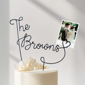 Könnte beinhalten: Ein Tortenaufsatz für eine Hochzeit mit den Worten "The Browns" in schwarzer Drahtschrift. Der Aufsatz enthält ein kleines Foto und weiße Blumen. Der Kuchen ist weiß.