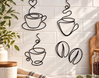 Puede incluir: Un conjunto de cuatro piezas de arte mural de metal negro con dibujos de línea continua de tres tazas de café diferentes y dos granos de café. Los diseños incluyen una taza con vapor en forma de corazón, una taza con vapor y una taza con asa.