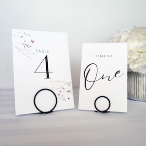 May include: Two white table number cards with black text and floral accents. One card reads "Table 4" and the other reads "Table No. One." Each card is held by a black metal ring stand. The cards are for a wedding or event.