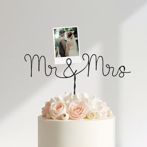 May include: A wedding cake topper featuring the words "Mr & Mrs" in black wire. A small photograph of a couple is attached. The cake is adorned with white and pink flowers.