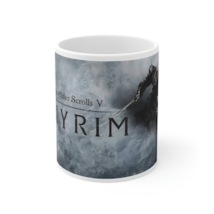 May include: White ceramic mug with a graphic of a character from the video game "The Elder Scrolls V: Skyrim". The character is holding a sword and is standing in front of a snowy mountain range. The text "Skyrim" is printed in black on the mug.