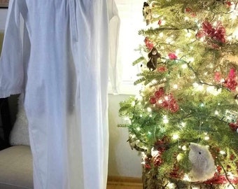 Puede incluir: Un camisón largo y blanco con mangas largas se exhibe en un maniquí. El camisón tiene una parte delantera abotonada y un cuello alto. Un árbol de Navidad decorado con luces y adornos está en el fondo.