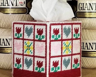 May include: A red, white, and blue tissue box cover with a patchwork design featuring tulips, hearts, and flowers. The cover is made of needlepoint and is sitting on a wooden shelf with balls of yarn.