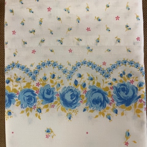 Könnte beinhalten: Weißer Baumwollstoff mit einem blauen und gelben Blumenmuster. Der Stoff hat ein kleines, verstreutes Blumenmuster und einen größeren Rand aus blauen Rosen mit gelben Blättern.