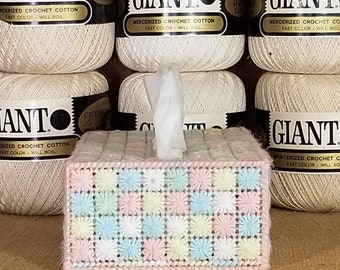 May include: A crocheted tissue box cover with a pink base and a checkered pattern of pastel blue, yellow, and pink squares. The cover is sitting on a brown woven surface.