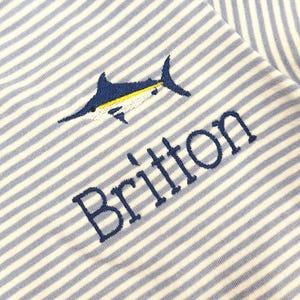 May include: A light blue and white striped t-shirt with a marlin fish embroidered on the front. The name "Britton" is embroidered below the fish. The shirt has a light blue collar and sleeve trim.
