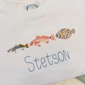 Op de afbeelding: Wit T-shirt met geborduurde vissen in blauw, oranje en bruin. De naam "Stetson" is eronder in blauwe letters geborduurd. Het shirt ligt op een gestreepte placemat.