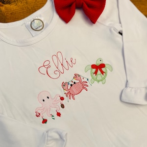 May include: White long-sleeved shirt with embroidered sea creatures and the name "Ellie" in red script. Features an octopus, crab, and turtle, each with festive embellishments. A large red bow sits at the top. The shirt has ruffled sleeves.