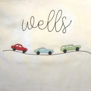 Puede incluir: Tela blanca con la palabra "wells" bordada en negro en cursiva. Debajo, tres coches bordados en rojo, azul claro y verde, cada uno con ruedas negras, están colocados sobre una línea negra.