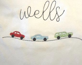 Puede incluir: Tela blanca con la palabra "wells" bordada en negro en cursiva. Debajo, tres coches bordados en rojo, azul claro y verde, cada uno con ruedas negras, están colocados sobre una línea negra.