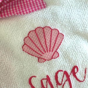 May include: White towel with a pink embroidered seashell design and the name "Sage" in cursive. A pink and white gingham bow is visible in the upper left corner. The towel is made of a soft, textured material.