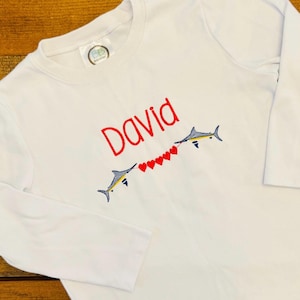May include: White long-sleeve shirt with the name "David" embroidered in red. Two stylized fish are embroidered below the name, connected by a string of red hearts. The fish are blue, yellow, and black.