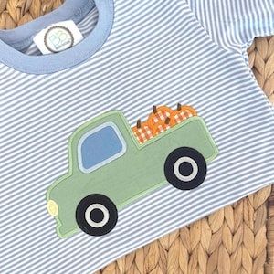 Puede incluir: Una camiseta de manga corta a rayas azul claro y blancas con cuello redondo. El centro presenta una aplicación de una camioneta verde salvia que transporta calabazas a cuadros naranjas y blancos. Se ve una etiqueta blanca con el texto BB Blanks Boutique.