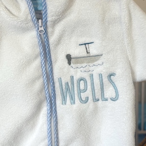 May include: White hooded terry cloth robe with a blue and white gingham trim. The robe features a boat design and the name "Wells" embroidered in light blue. It has a full zipper closure.