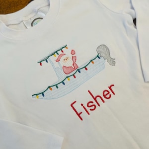 May include: White long-sleeved shirt with an embroidered design of Santa Claus in a boat decorated with colorful lights. The name "Fisher" is embroidered in red below the design. The shirt is made of a soft, white fabric.
