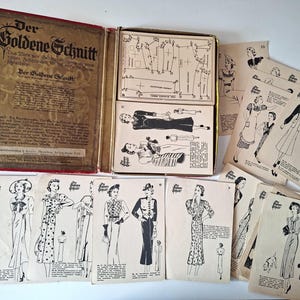 May include: A vintage Der Goldene Schnitt sewing pattern set featuring a red-bound instructional book, a metal storage box containing technical pattern sheets, and numerous individual paper cards displaying black and white fashion illustrations.