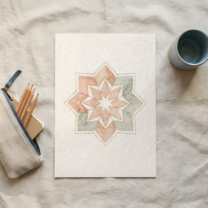 May include: Watercolor painting of a geometric starburst design in shades of peach, green, and gray. A light blue ceramic cup, a pencil case with pencils, and a smartphone are also visible.