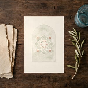 May include: A watercolor print of an arched window with a geometric star design in shades of green, pink, and white. The print is on a white card, next to a stack of handmade paper and a sprig of olive branches. A blue glass is also present.